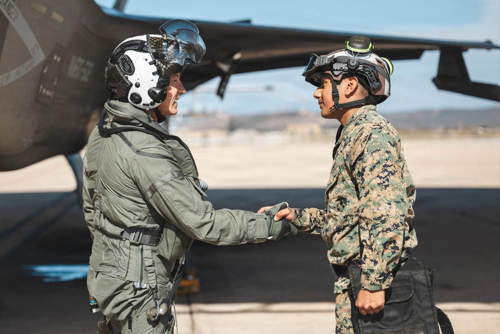 Two military personnel are shaking hands on an airfield tarmac in front of a military aircraft. One individual is wearing an olive green flight suit with a white pilot helmet equipped with a visor, while the other is wearing digital camouflage (MARPAT pattern) consistent with U.S. Marine Corps uniform and a tactical helmet. Both appear to be smiling, suggesting a positive interaction such as a post-mission debrief or greeting.