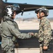 Two military personnel are shaking hands on an airfield tarmac in front of a military aircraft. One individual is wearing an olive green flight suit with a white pilot helmet equipped with a visor, while the other is wearing digital camouflage (MARPAT pattern) consistent with U.S. Marine Corps uniform and a tactical helmet. Both appear to be smiling, suggesting a positive interaction such as a post-mission debrief or greeting.