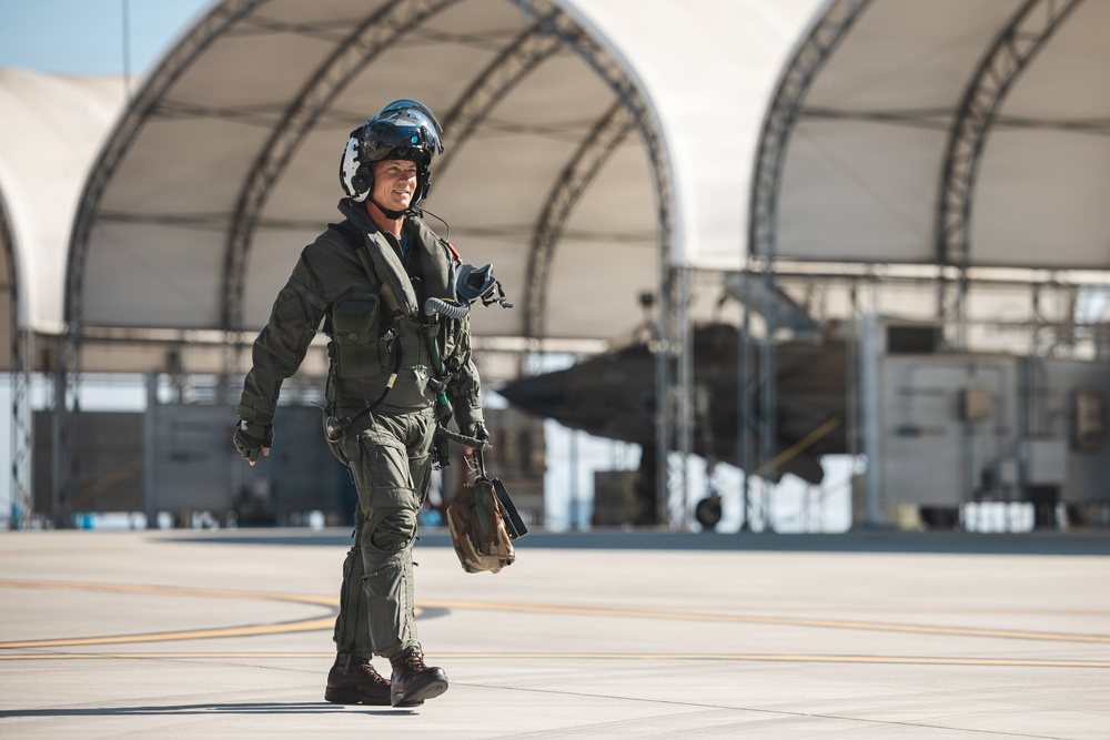 A military pilot in full flight gear walks across an airfield tarmac, smiling and appearing confident. The pilot is wearing an olive-green flight suit, helmet with visor, oxygen mask, and carrying flight equipment. In the background, large arched aircraft hangars are visible along with what appears to be a military jet aircraft.