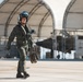 A military pilot in full flight gear walks across an airfield tarmac, smiling and appearing confident. The pilot is wearing an olive-green flight suit, helmet with visor, oxygen mask, and carrying flight equipment. In the background, large arched aircraft hangars are visible along with what appears to be a military jet aircraft.