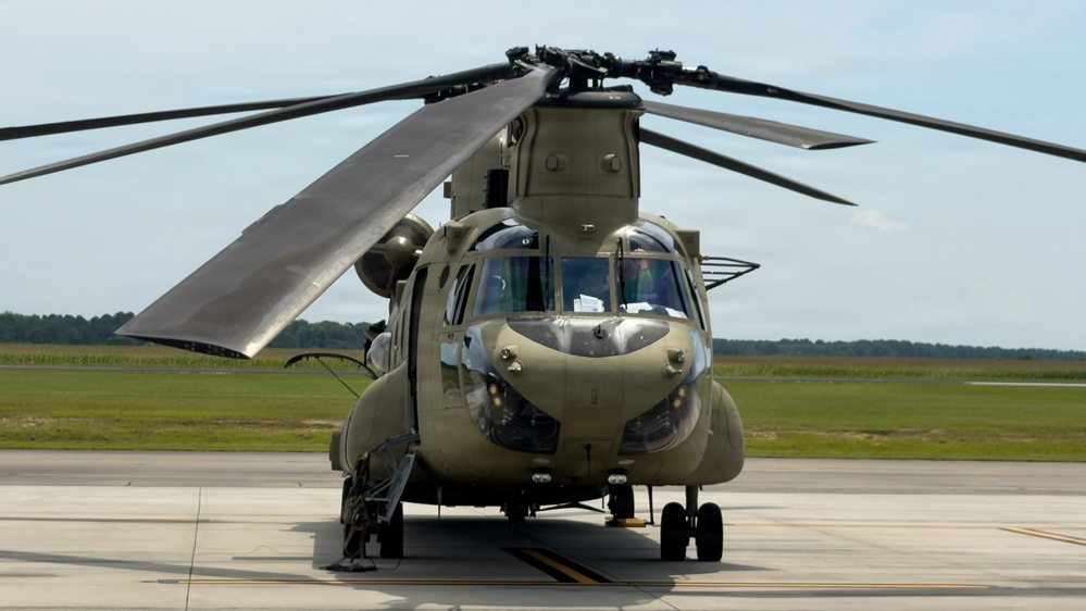 A military tandem-rotor helicopter, identifiable as a Boeing CH-47 Chinook, is parked on a concrete tarmac or airfield. The aircraft is painted in olive drab/army green and appears to be stationary with rotor blades extended. The setting appears to be a military airfield with flat grassy terrain and tree line visible in the background.
