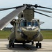 A military tandem-rotor helicopter, identifiable as a Boeing CH-47 Chinook, is parked on a concrete tarmac or airfield. The aircraft is painted in olive drab/army green and appears to be stationary with rotor blades extended. The setting appears to be a military airfield with flat grassy terrain and tree line visible in the background.