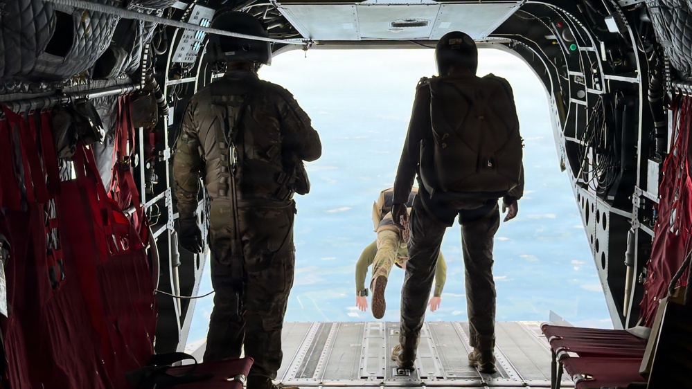 The image shows the interior of a military cargo aircraft with its rear ramp open during a parachute operation. Two personnel in military gear are standing at the ramp's edge while a third individual has already jumped and is visible in freefall just outside the aircraft. The ground and sky are visible through the open ramp, indicating significant altitude.