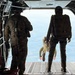 The image shows the interior of a military cargo aircraft with its rear ramp open during a parachute operation. Two personnel in military gear are standing at the ramp's edge while a third individual has already jumped and is visible in freefall just outside the aircraft. The ground and sky are visible through the open ramp, indicating significant altitude.