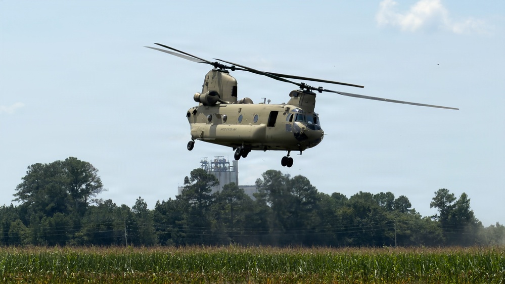 A U.S. Army CH-47 Chinook tandem-rotor helicopter is captured in low-level flight over an agricultural field, likely a cornfield. The aircraft is painted in standard Army tan/desert camouflage and appears to be either landing or conducting a low-altitude pass. The background features a treeline, power lines, and what appears to be an industrial or grain storage facility.