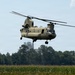 A U.S. Army CH-47 Chinook tandem-rotor helicopter is captured in low-level flight over an agricultural field, likely a cornfield. The aircraft is painted in standard Army tan/desert camouflage and appears to be either landing or conducting a low-altitude pass. The background features a treeline, power lines, and what appears to be an industrial or grain storage facility.