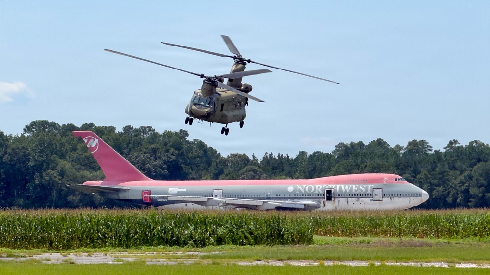 A military CH-47 Chinook tandem-rotor helicopter is flying low over a field near a stationary Northwest Airlines Boeing 747 wide-body jet. The 747 appears to be parked or decommissioned on a grass/tarmac area surrounded by what looks like a cornfield and tree line. The color scheme of the Northwest Airlines aircraft shows its distinctive red and gray livery.