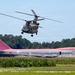 A military CH-47 Chinook tandem-rotor helicopter is flying low over a field near a stationary Northwest Airlines Boeing 747 wide-body jet. The 747 appears to be parked or decommissioned on a grass/tarmac area surrounded by what looks like a cornfield and tree line. The color scheme of the Northwest Airlines aircraft shows its distinctive red and gray livery.