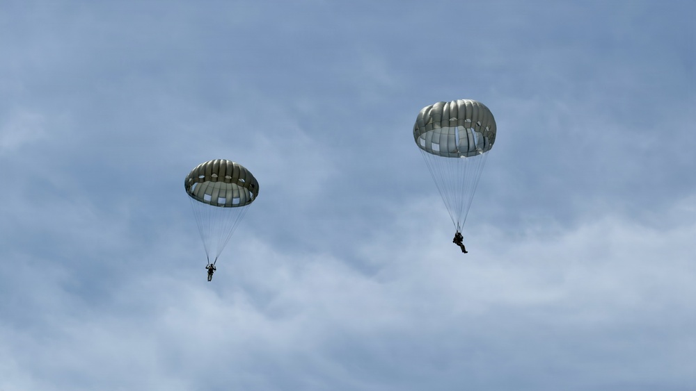 Two military paratroopers are descending through a partly cloudy sky using round, olive-drab/gray parachutes typical of military static-line deployments. Both canopies are fully deployed and appear to be in stable descent. The image was taken from a ground-level perspective looking upward.