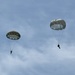 Two military paratroopers are descending through a partly cloudy sky using round, olive-drab/gray parachutes typical of military static-line deployments. Both canopies are fully deployed and appear to be in stable descent. The image was taken from a ground-level perspective looking upward.