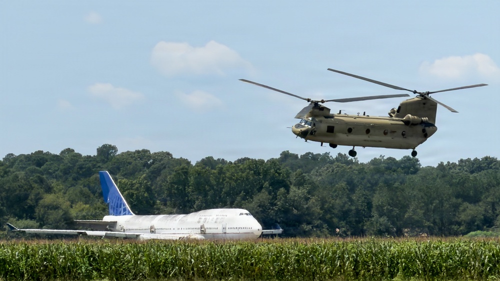 A military CH-47 Chinook tandem-rotor helicopter in olive drab livery is airborne over a rural area with cornfields in the foreground. In the background, a large white commercial Boeing 747 aircraft with a blue tail bearing a partial airline logo is parked or stationary on the ground. The scene appears to be near a small airfield or open rural area bordered by dense tree lines.
