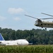 A military CH-47 Chinook tandem-rotor helicopter in olive drab livery is airborne over a rural area with cornfields in the foreground. In the background, a large white commercial Boeing 747 aircraft with a blue tail bearing a partial airline logo is parked or stationary on the ground. The scene appears to be near a small airfield or open rural area bordered by dense tree lines.