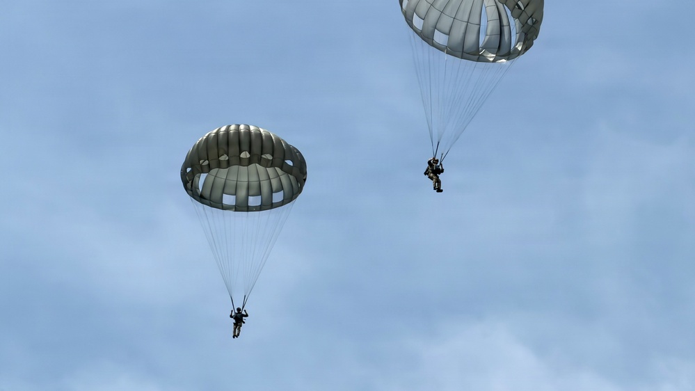 The image shows two military paratroopers descending under round, olive-drab/gray parachutes against a partly cloudy blue sky. Both parachutes appear to be standard military static-line round canopies, consistent with airborne operations. The paratroopers are visible suspended beneath their respective canopies at different altitudes.