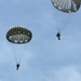 The image shows two military paratroopers descending under round, olive-drab/gray parachutes against a partly cloudy blue sky. Both parachutes appear to be standard military static-line round canopies, consistent with airborne operations. The paratroopers are visible suspended beneath their respective canopies at different altitudes.