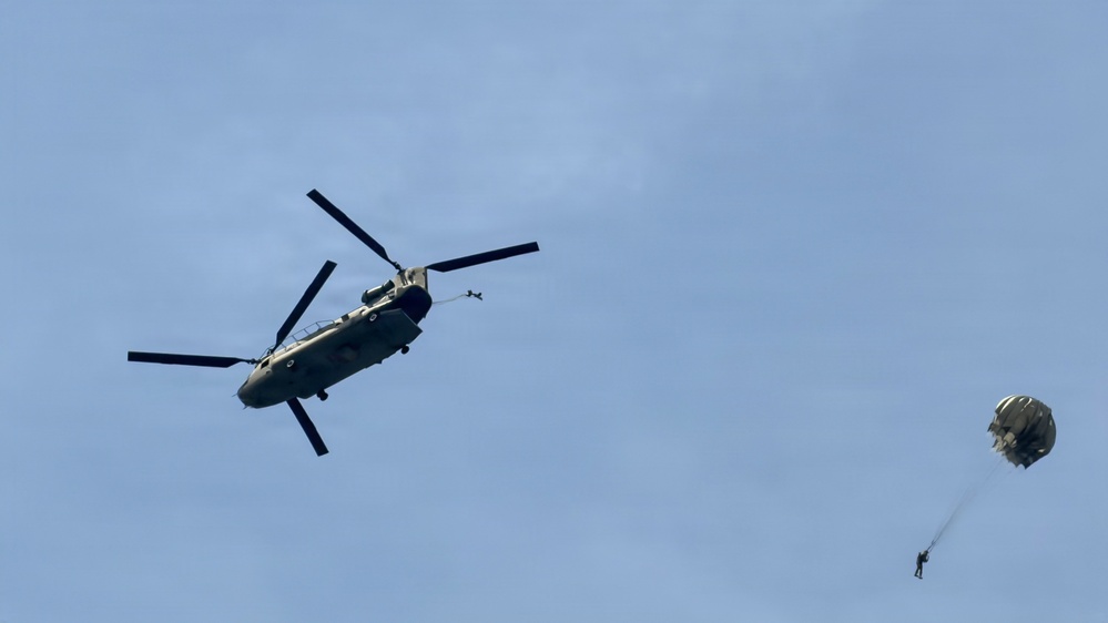 A military tandem-rotor helicopter, consistent with a CH-47 Chinook, is captured in flight against a pale blue sky. To the right of the helicopter, a parachutist is visible descending with a partially deployed or round parachute canopy above them. This appears to be a military airborne operation or training exercise involving a helicopter jump.
