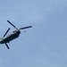 A military tandem-rotor helicopter, consistent with a CH-47 Chinook, is captured in flight against a pale blue sky. To the right of the helicopter, a parachutist is visible descending with a partially deployed or round parachute canopy above them. This appears to be a military airborne operation or training exercise involving a helicopter jump.