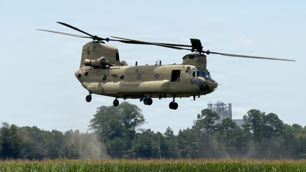 A United States Army CH-47 Chinook tandem-rotor helicopter is hovering at low altitude over a cornfield, with rotor wash visibly kicking up dust and debris near the tree line. The aircraft is painted in a tan/desert camouflage scheme and bears 'UNITED STATES ARMY' markings on its fuselage. The setting appears to be a rural agricultural area with trees and an industrial structure visible in the background.