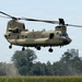 A United States Army CH-47 Chinook tandem-rotor helicopter is hovering at low altitude over a cornfield, with rotor wash visibly kicking up dust and debris near the tree line. The aircraft is painted in a tan/desert camouflage scheme and bears 'UNITED STATES ARMY' markings on its fuselage. The setting appears to be a rural agricultural area with trees and an industrial structure visible in the background.