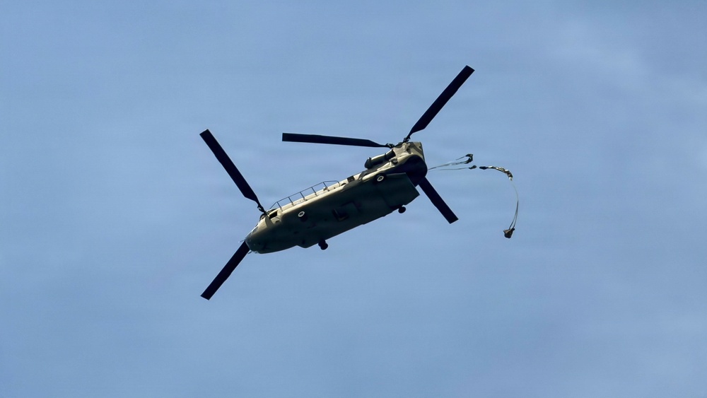 A military tandem-rotor helicopter, identifiable as a Boeing CH-47 Chinook, is photographed in flight against a clear blue sky. The helicopter appears to be in a slightly banked orientation, and a line or cable with what appears to be a small object or parachute is trailing from its rear. The aircraft displays an olive drab/military green color scheme consistent with military service.