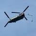 A military tandem-rotor helicopter, identifiable as a Boeing CH-47 Chinook, is photographed in flight against a clear blue sky. The helicopter appears to be in a slightly banked orientation, and a line or cable with what appears to be a small object or parachute is trailing from its rear. The aircraft displays an olive drab/military green color scheme consistent with military service.