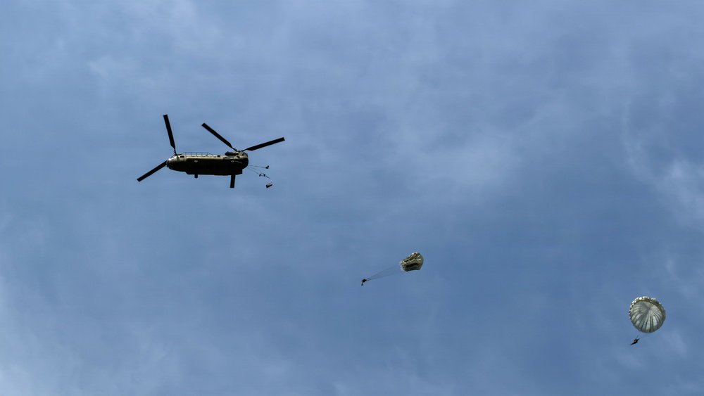 A military tandem-rotor helicopter, consistent with a CH-47 Chinook, is conducting an airborne drop operation at altitude against a partly cloudy blue sky. Two parachutes are visible in various stages of deployment to the right of the helicopter, with personnel or cargo suspended beneath them. This appears to be a military parachute training or operational insertion exercise.