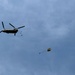 A military tandem-rotor helicopter, consistent with a CH-47 Chinook, is conducting an airborne drop operation at altitude against a partly cloudy blue sky. Two parachutes are visible in various stages of deployment to the right of the helicopter, with personnel or cargo suspended beneath them. This appears to be a military parachute training or operational insertion exercise.