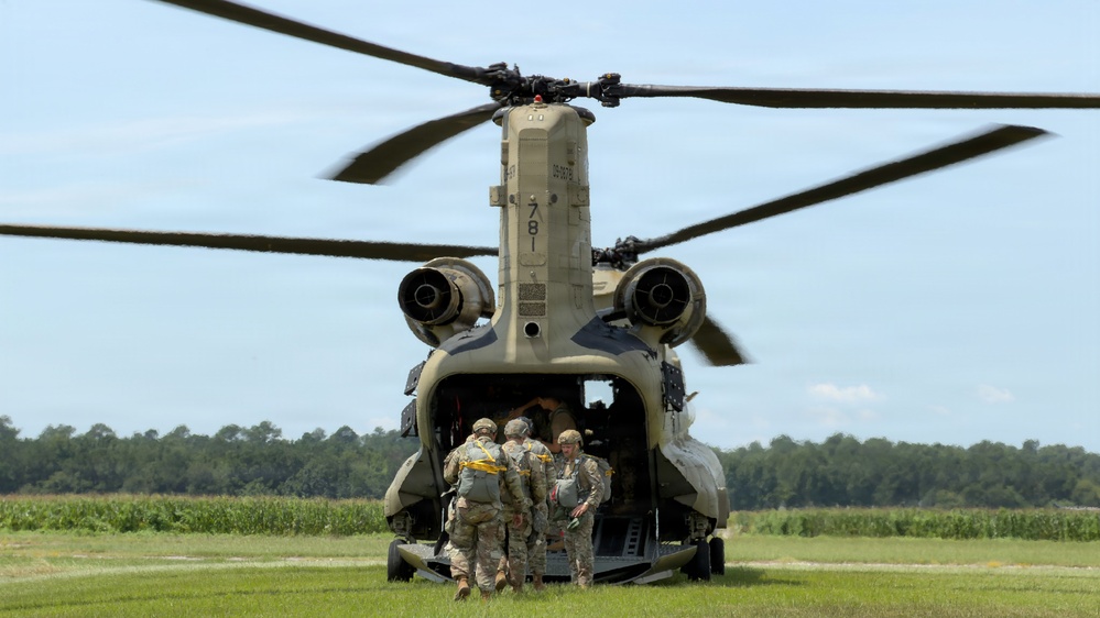 A Boeing CH-47 Chinook tandem-rotor military helicopter, tail number 781, is on the ground with its rear ramp lowered. Several soldiers in camouflage uniforms and carrying equipment are boarding or preparing to board through the aft ramp. The scene appears to be a training or operational exercise at a grassy field with tree line and crop fields visible in the background.