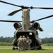 A Boeing CH-47 Chinook tandem-rotor military helicopter, tail number 781, is on the ground with its rear ramp lowered. Several soldiers in camouflage uniforms and carrying equipment are boarding or preparing to board through the aft ramp. The scene appears to be a training or operational exercise at a grassy field with tree line and crop fields visible in the background.