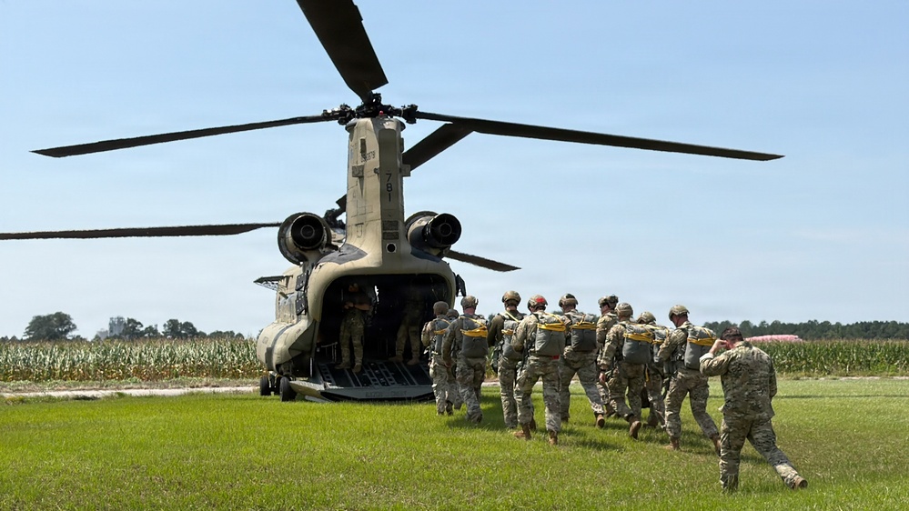 A line of military personnel in full combat gear and parachute equipment are boarding a CH-47 Chinook helicopter via its rear ramp. The operation appears to be a airborne/parachute insertion exercise taking place on a grassy field adjacent to a cornfield. The helicopter bears the tail number '781' and is in a landed, rotors-spinning configuration.
