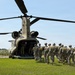 A line of military personnel in full combat gear and parachute equipment are boarding a CH-47 Chinook helicopter via its rear ramp. The operation appears to be a airborne/parachute insertion exercise taking place on a grassy field adjacent to a cornfield. The helicopter bears the tail number '781' and is in a landed, rotors-spinning configuration.