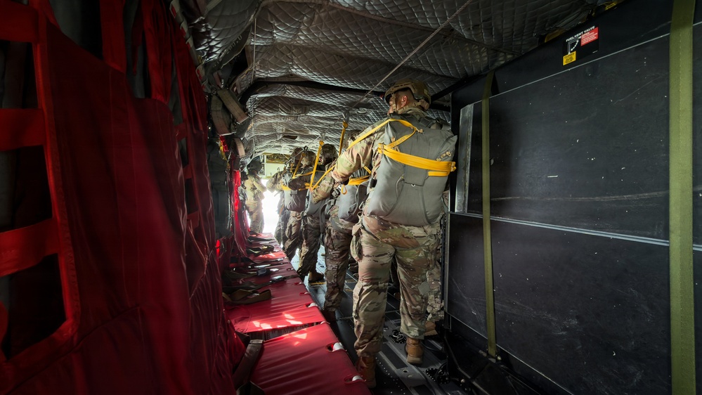 A line of military paratroopers stands inside a military transport aircraft, equipped with parachute rigs and helmets, preparing for a jump. The personnel are lined up along the interior of the aircraft facing toward the open rear ramp, with static lines attached overhead. The interior features characteristic red canvas seating, quilted insulation panels on the ceiling, and yellow anchor line cables.
