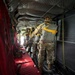 A line of military paratroopers stands inside a military transport aircraft, equipped with parachute rigs and helmets, preparing for a jump. The personnel are lined up along the interior of the aircraft facing toward the open rear ramp, with static lines attached overhead. The interior features characteristic red canvas seating, quilted insulation panels on the ceiling, and yellow anchor line cables.