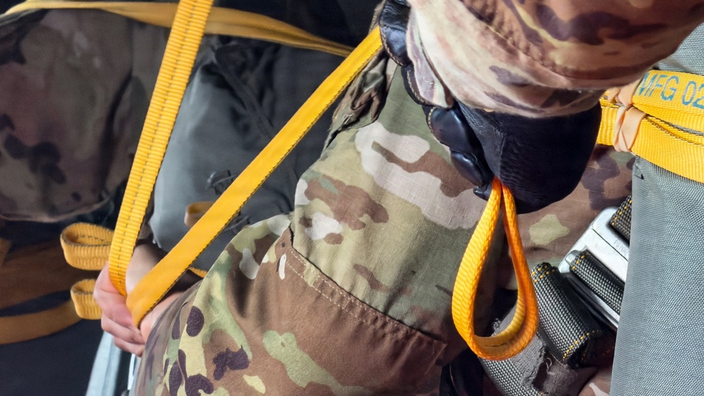 A close-up image of a person wearing MultiCam/OCP camouflage military uniform with yellow/gold nylon straps crossing their torso and midsection. The straps appear to be part of a harness or restraint system, with visible hardware including what appears to be a buckle or connector on the right side. A partial text label reading 'MFG 02' is visible on one of the yellow straps.