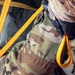 A close-up image of a person wearing MultiCam/OCP camouflage military uniform with yellow/gold nylon straps crossing their torso and midsection. The straps appear to be part of a harness or restraint system, with visible hardware including what appears to be a buckle or connector on the right side. A partial text label reading 'MFG 02' is visible on one of the yellow straps.