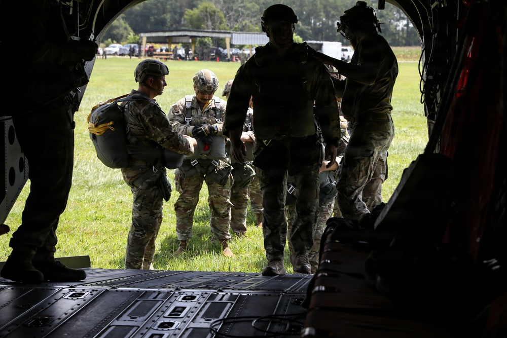 Military personnel in multicam/OCP camouflage uniforms are gathered at the rear ramp of what appears to be a CH-47 Chinook helicopter on a grassy field. The soldiers are equipped with parachute rigs and tactical helmets, suggesting pre-jump preparation or equipment checks. The scene appears to be a airborne operation or training exercise at a military installation.