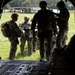 Military personnel in multicam/OCP camouflage uniforms are gathered at the rear ramp of what appears to be a CH-47 Chinook helicopter on a grassy field. The soldiers are equipped with parachute rigs and tactical helmets, suggesting pre-jump preparation or equipment checks. The scene appears to be a airborne operation or training exercise at a military installation.