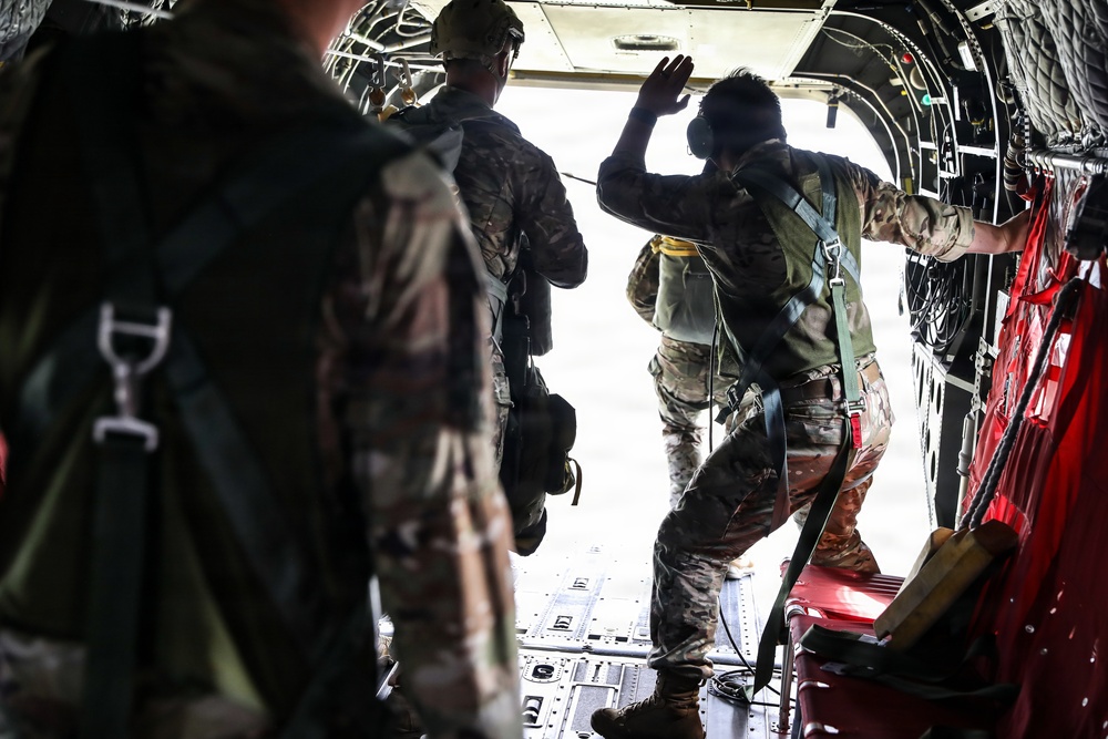 Military personnel in camouflage uniforms are positioned near the open rear ramp of a military transport aircraft, appearing to prepare for a parachute jump operation. One individual near the opening is wearing an oxygen/breathing mask and has a hand raised, possibly signaling. The interior of the aircraft shows red seating, static lines, and standard military cargo aircraft features.