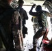 Military personnel in camouflage uniforms are positioned near the open rear ramp of a military transport aircraft, appearing to prepare for a parachute jump operation. One individual near the opening is wearing an oxygen/breathing mask and has a hand raised, possibly signaling. The interior of the aircraft shows red seating, static lines, and standard military cargo aircraft features.