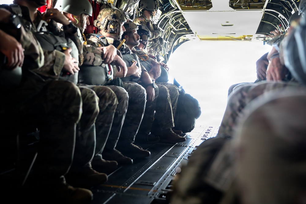 A group of military personnel in full combat gear are seated along the interior wall of a military transport aircraft, likely a C-130 or similar cargo plane, with the rear ramp open. The soldiers appear to be preparing for a parachute jump or airborne insertion operation, as indicated by their helmets, tactical gear, and positioning near the open ramp. The bright light from the open ramp suggests the aircraft is airborne.