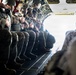 A group of military personnel in full combat gear are seated along the interior wall of a military transport aircraft, likely a C-130 or similar cargo plane, with the rear ramp open. The soldiers appear to be preparing for a parachute jump or airborne insertion operation, as indicated by their helmets, tactical gear, and positioning near the open ramp. The bright light from the open ramp suggests the aircraft is airborne.