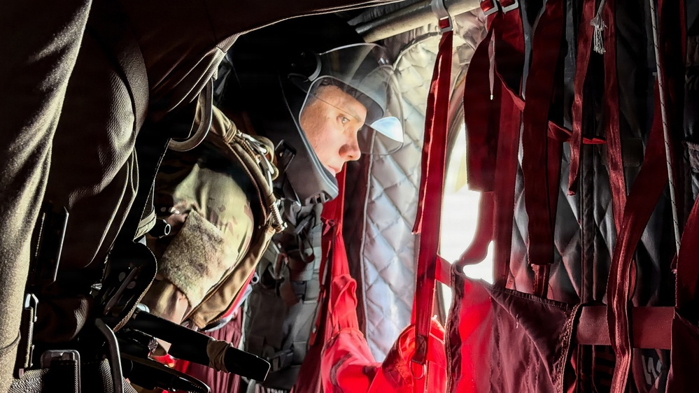 A military personnel member is seated inside what appears to be a military helicopter or transport aircraft, wearing tactical gear including a helmet and body armor. The interior shows characteristic red cargo straps and quilted insulation panels typical of military rotary-wing aircraft. The individual appears to be looking out through a window or opening, with dramatic lighting contrasting the dark interior.