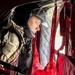 A military personnel member is seated inside what appears to be a military helicopter or transport aircraft, wearing tactical gear including a helmet and body armor. The interior shows characteristic red cargo straps and quilted insulation panels typical of military rotary-wing aircraft. The individual appears to be looking out through a window or opening, with dramatic lighting contrasting the dark interior.