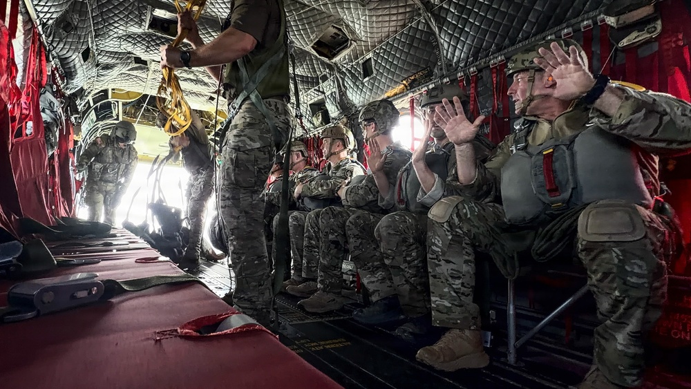 Military personnel in full combat gear and parachute equipment are seated inside a military transport aircraft, appearing to be preparing for a parachute jump. The aircraft's rear ramp is open, revealing bright light outside, while a jumpmaster stands near the opening holding static lines. The soldiers are equipped with helmets, tactical vests, and parachute harnesses, suggesting an airborne operation or training exercise.