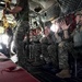 Military personnel in full combat gear and parachute equipment are seated inside a military transport aircraft, appearing to be preparing for a parachute jump. The aircraft's rear ramp is open, revealing bright light outside, while a jumpmaster stands near the opening holding static lines. The soldiers are equipped with helmets, tactical vests, and parachute harnesses, suggesting an airborne operation or training exercise.