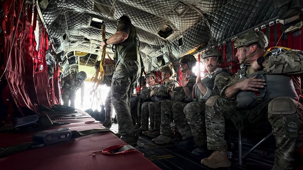 Military personnel are seated inside a large military transport aircraft, appearing to be preparing for a parachute jump operation. A standing crew member or jumpmaster is managing static lines near the open rear of the aircraft. The soldiers are equipped with combat gear, helmets, and parachute equipment.