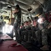 Military personnel are seated inside a large military transport aircraft, appearing to be preparing for a parachute jump operation. A standing crew member or jumpmaster is managing static lines near the open rear of the aircraft. The soldiers are equipped with combat gear, helmets, and parachute equipment.