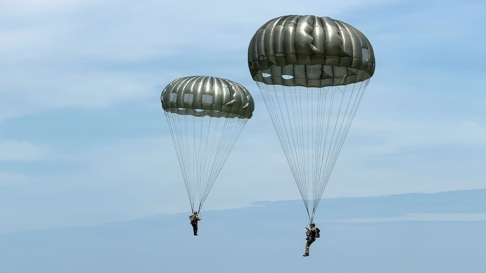 Two military paratroopers are descending under fully deployed olive-drab/silver round parachutes against a partly cloudy blue sky. The parachutes appear to be standard military static-line round canopies, consistent with airborne operations. Both jumpers are visible beneath their respective canopies with full military gear.