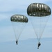 Two military paratroopers are descending under fully deployed olive-drab/silver round parachutes against a partly cloudy blue sky. The parachutes appear to be standard military static-line round canopies, consistent with airborne operations. Both jumpers are visible beneath their respective canopies with full military gear.