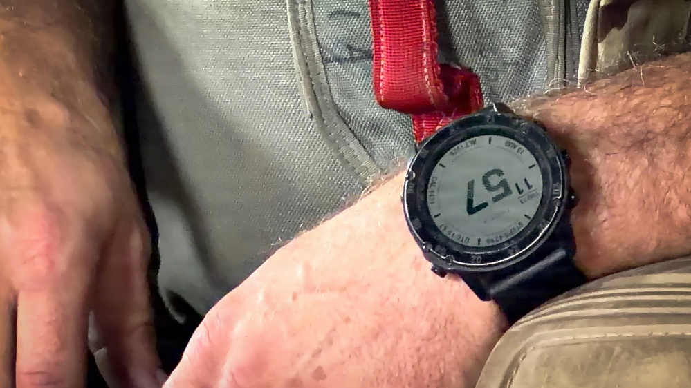 A close-up image of a digital smartwatch worn on a person's wrist, appearing to be a Garmin-style GPS/sport watch with a dark bezel and black band. The watch display shows the time as 11:57, along with additional data fields typical of a multisport or tactical watch. The wearer appears to be seated, with khaki/olive clothing and a red strap visible in the background.
