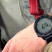 A close-up image of a digital smartwatch worn on a person's wrist, appearing to be a Garmin-style GPS/sport watch with a dark bezel and black band. The watch display shows the time as 11:57, along with additional data fields typical of a multisport or tactical watch. The wearer appears to be seated, with khaki/olive clothing and a red strap visible in the background.